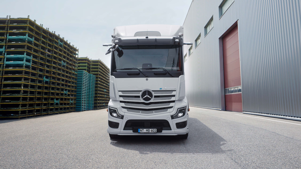 Ein eActros 600 von Mercedes-Benz Trucks steht frontal auf einer asphaltierten Fläche vor einem modernen Gebäude mit einer langen Außentreppe. Der Lkw ist weiß und hat das Mercedes-Stern-Logo auf dem Kühlergrill, während im Hintergrund Bäume und weitere Gebäudestrukturen zu sehen sind.