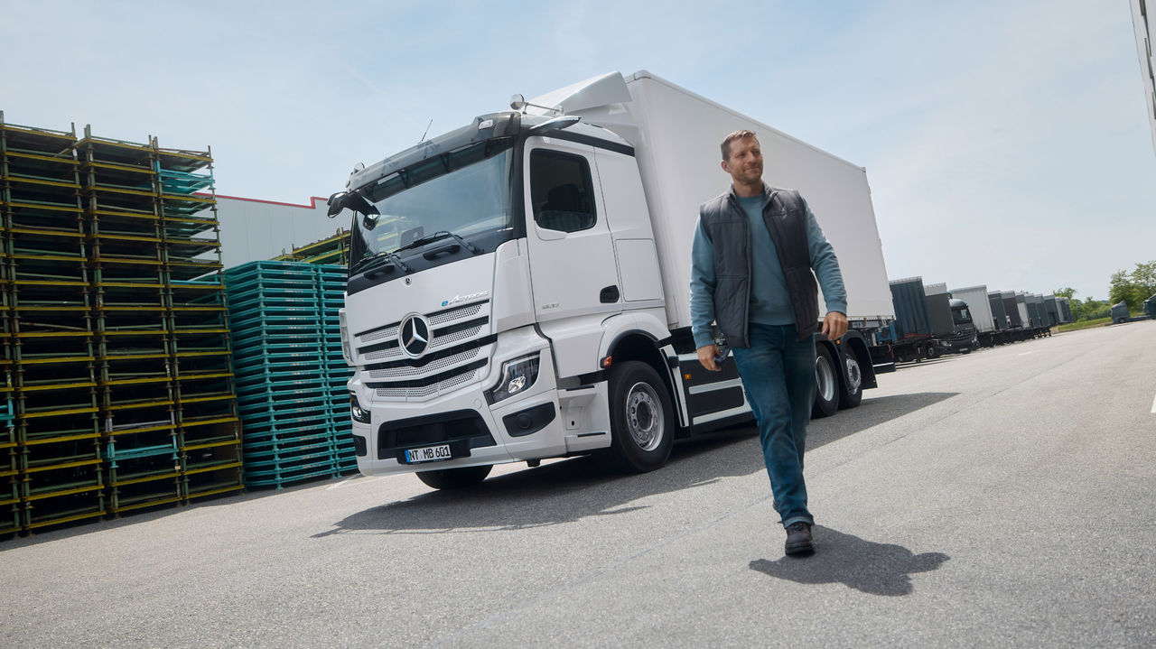 Ein Mann in Arbeitskleidung geht auf einer asphaltierten Fläche, während im Hintergrund ein weißer eActros 600 von Mercedes-Benz Trucks mit einem Anhänger parkt. Links sind gestapelte Paletten zu sehen und rechts ein großes Gebäude, das auf einen Lager- oder Industriekomplex hindeutet.